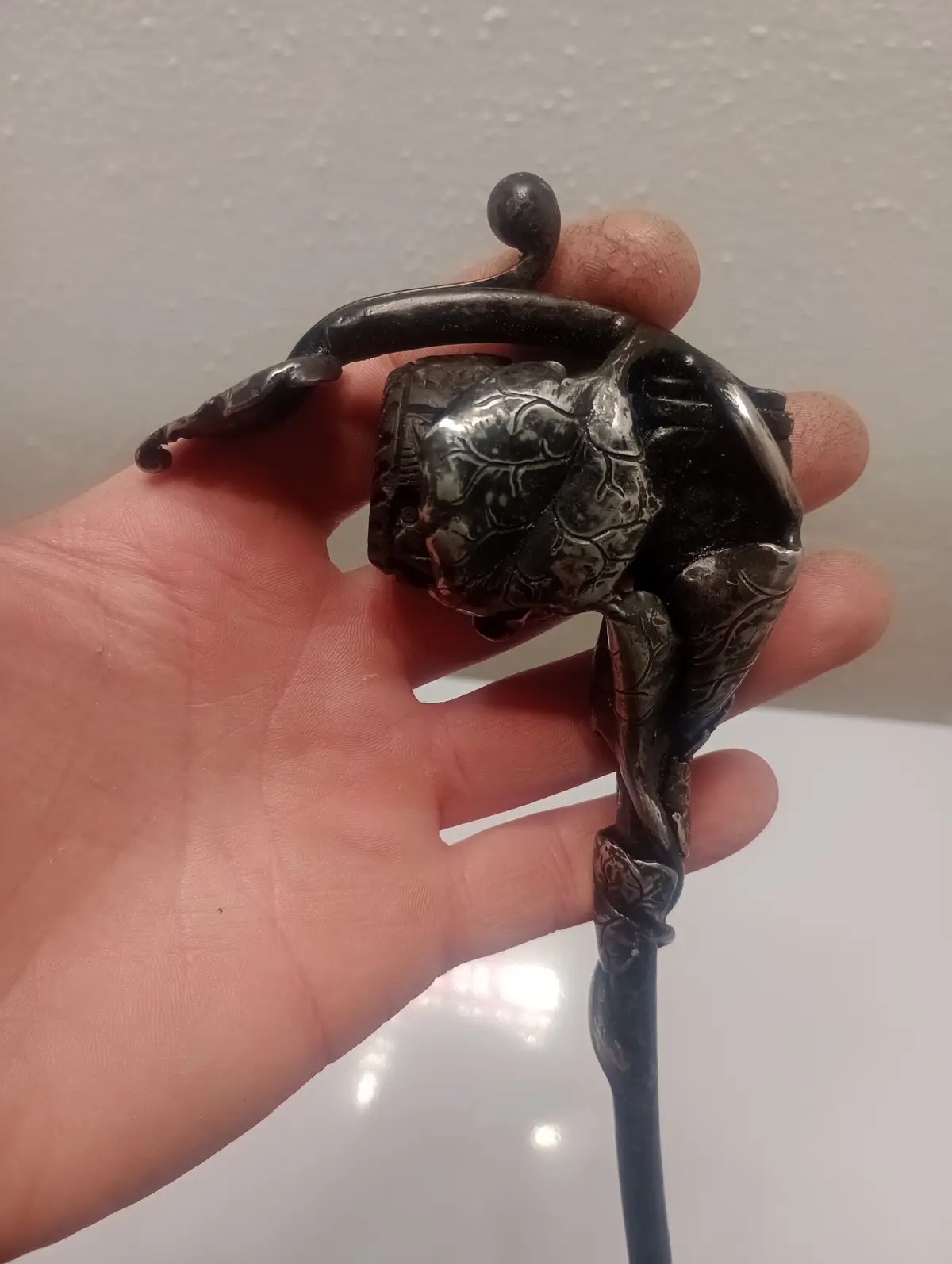 A hand holds a small, dark, ornate figurine that appears to be a stylized human or mythical creature in a contorted pose…