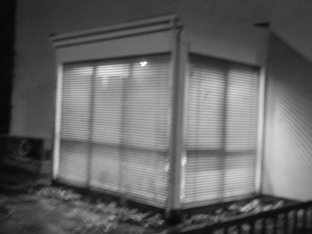 A blurry black-and-white photo shows two windows with horizontal blinds, one of which has a small light visible through the…