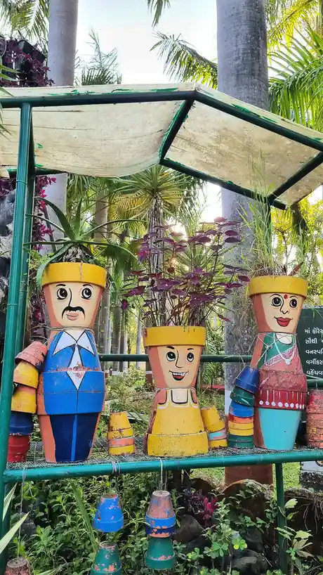 Three whimsical, hand-painted figures made from stacked terracotta pots stand under a green-framed canopy in a lush garden…