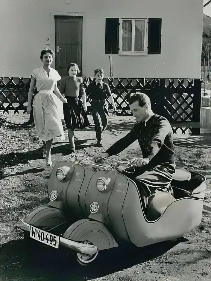 A vintage black-and-white photo shows a man sitting on a scooter with a woman and two children walking behind him. The man…