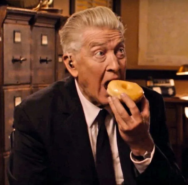 An older man in a suit and tie is shown mid-bite into a glazed donut, his eyes wide open in exaggerated shock or surprise…
