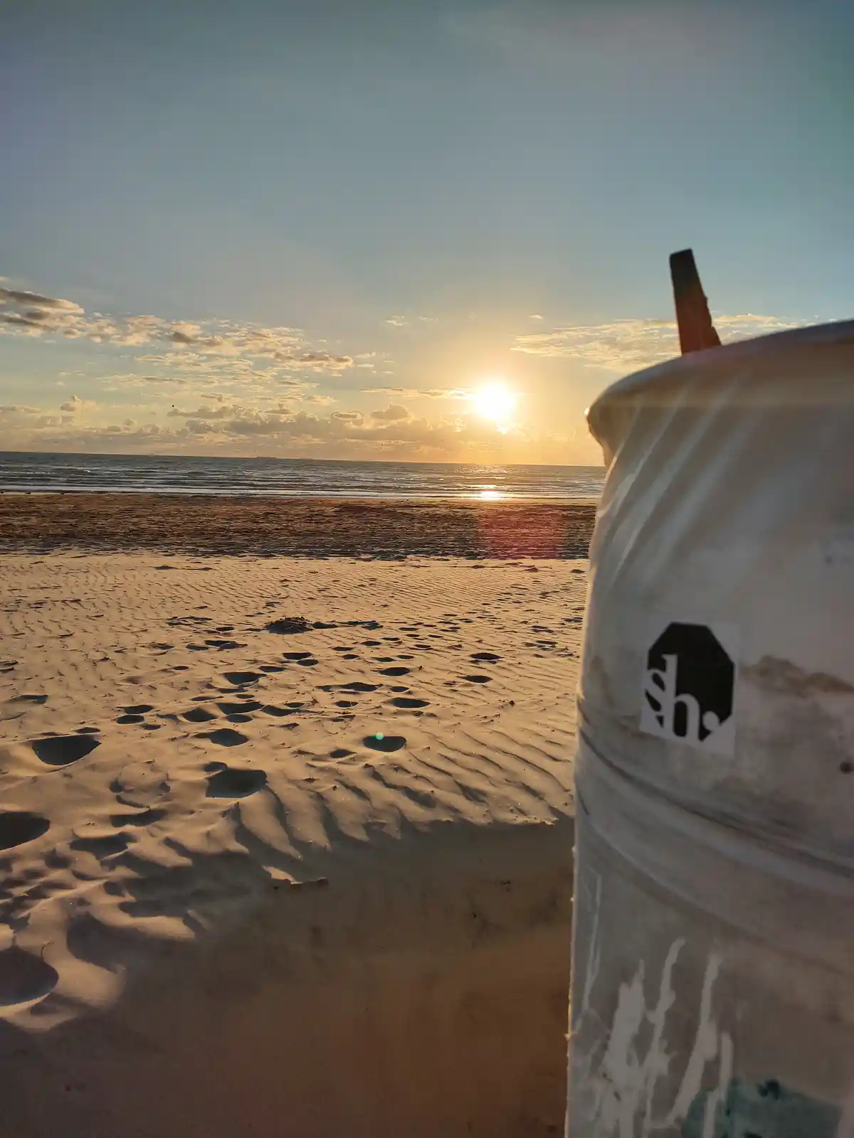 A serene beach sunset with the sun low on the horizon over the ocean, casting golden light on rippled sand with footprints…