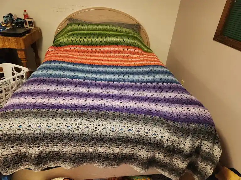 A bed is covered with a large, hand-crocheted blanket featuring wide horizontal stripes that transition through a rainbow of…