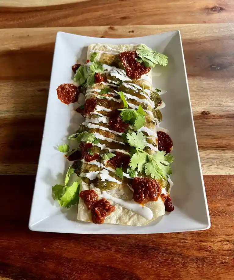 A long, rolled burrito sits on a white rectangular plate, garnished with green cilantro leaves, a drizzle of white sauce…
