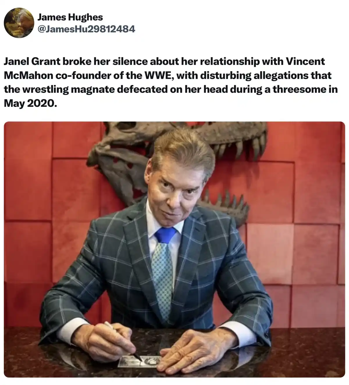 A man in a plaid suit and blue tie sits at a dark desk, signing a document. Behind him, a large dinosaur skull is mounted on…