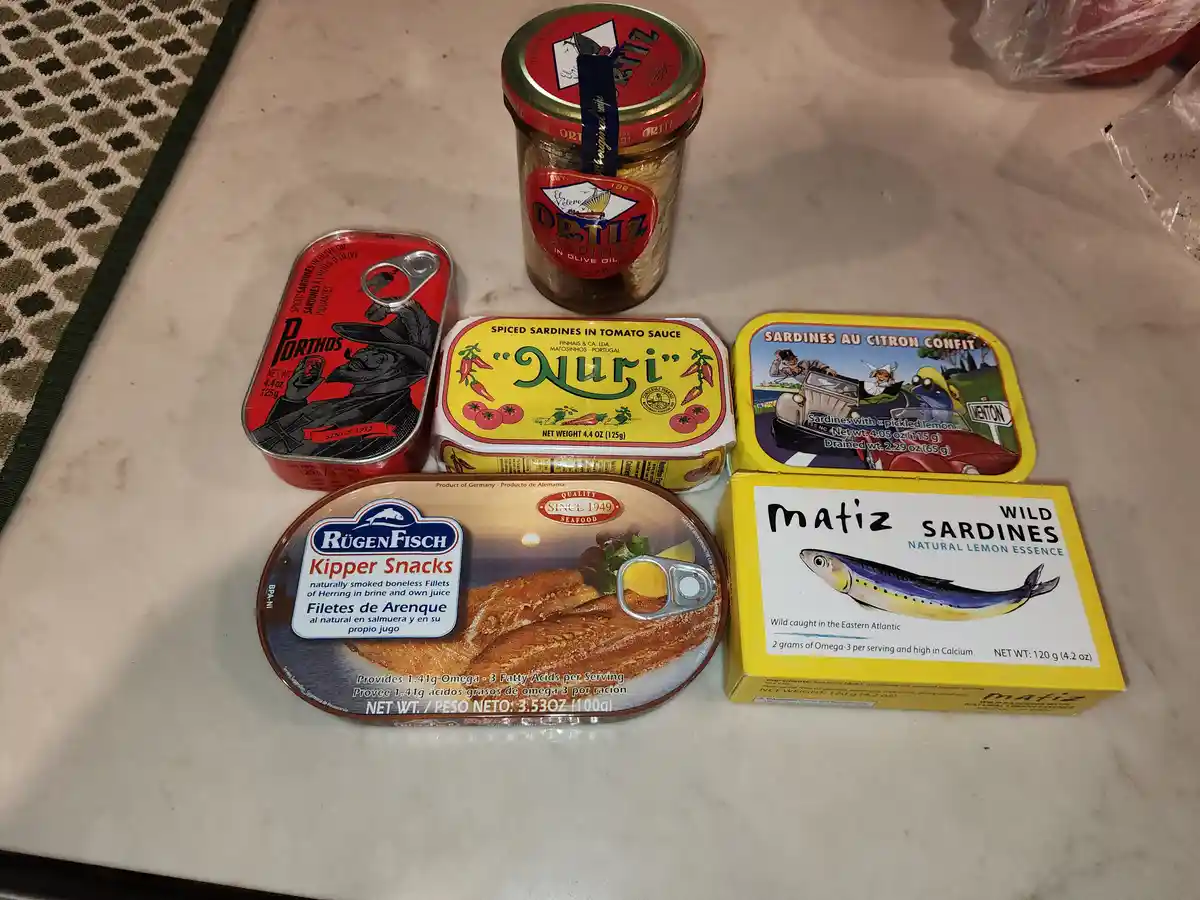 A collection of six different canned and jarred fish products, including sardines and herring, arranged on a white…