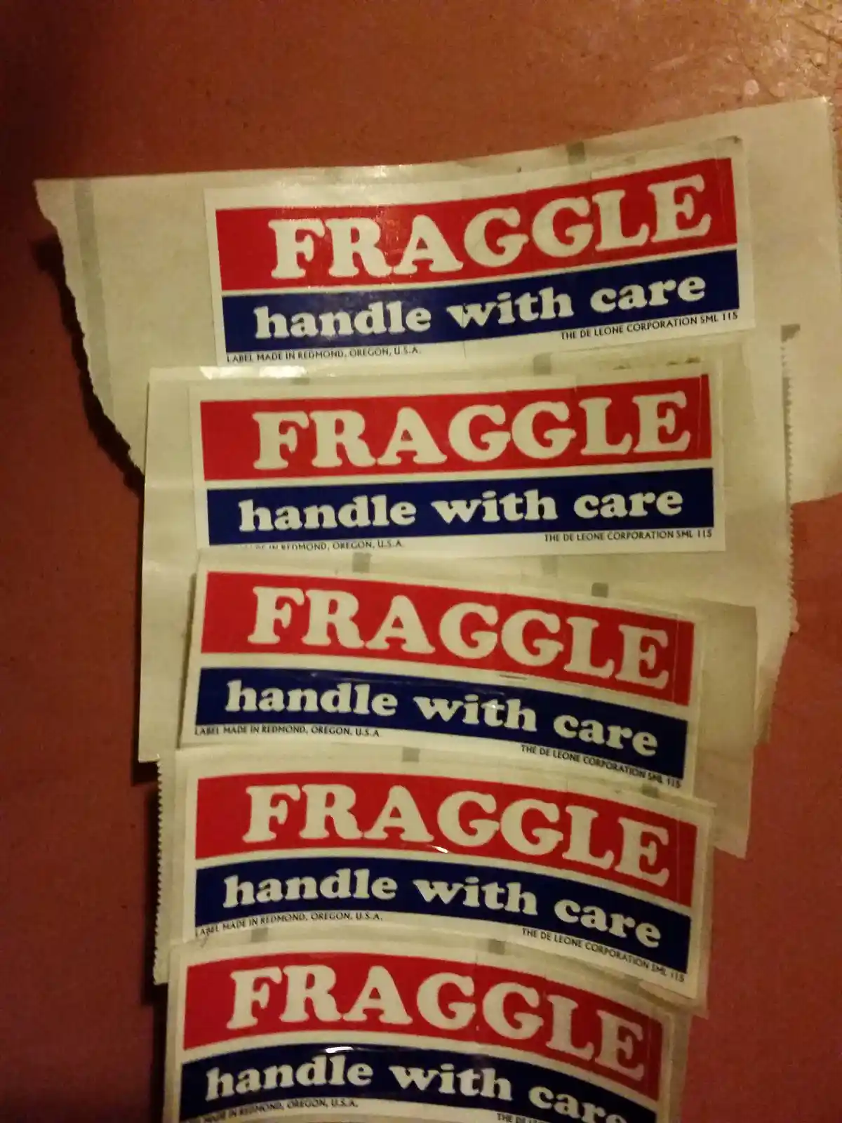 Several identical 'FRAGGLE handle with care' shipping labels are stacked vertically on a reddish-brown surface. Each label…