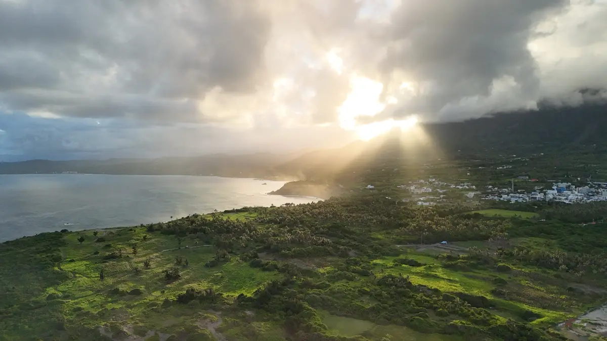 An aerial view captures sunlight breaking through heavy clouds over a lush green coastal landscape with rolling hills, a…