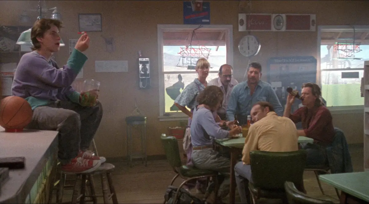 A young man in a purple sweater sits on a stool at a diner counter, holding a red marker and a cup of candy, with a…