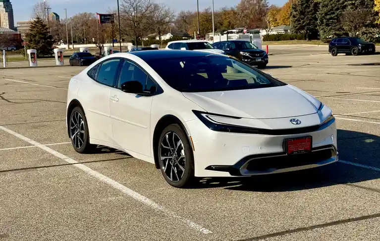 A white Toyota Prius is parked in a lot with other cars and charging stations visible in the background. The car has a…