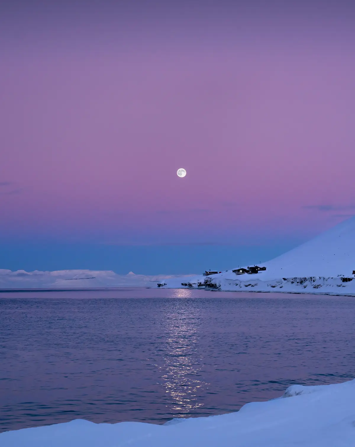 A serene, snowy landscape at twilight with a full moon reflecting on calm water, small cabins nestled on a snow-covered…