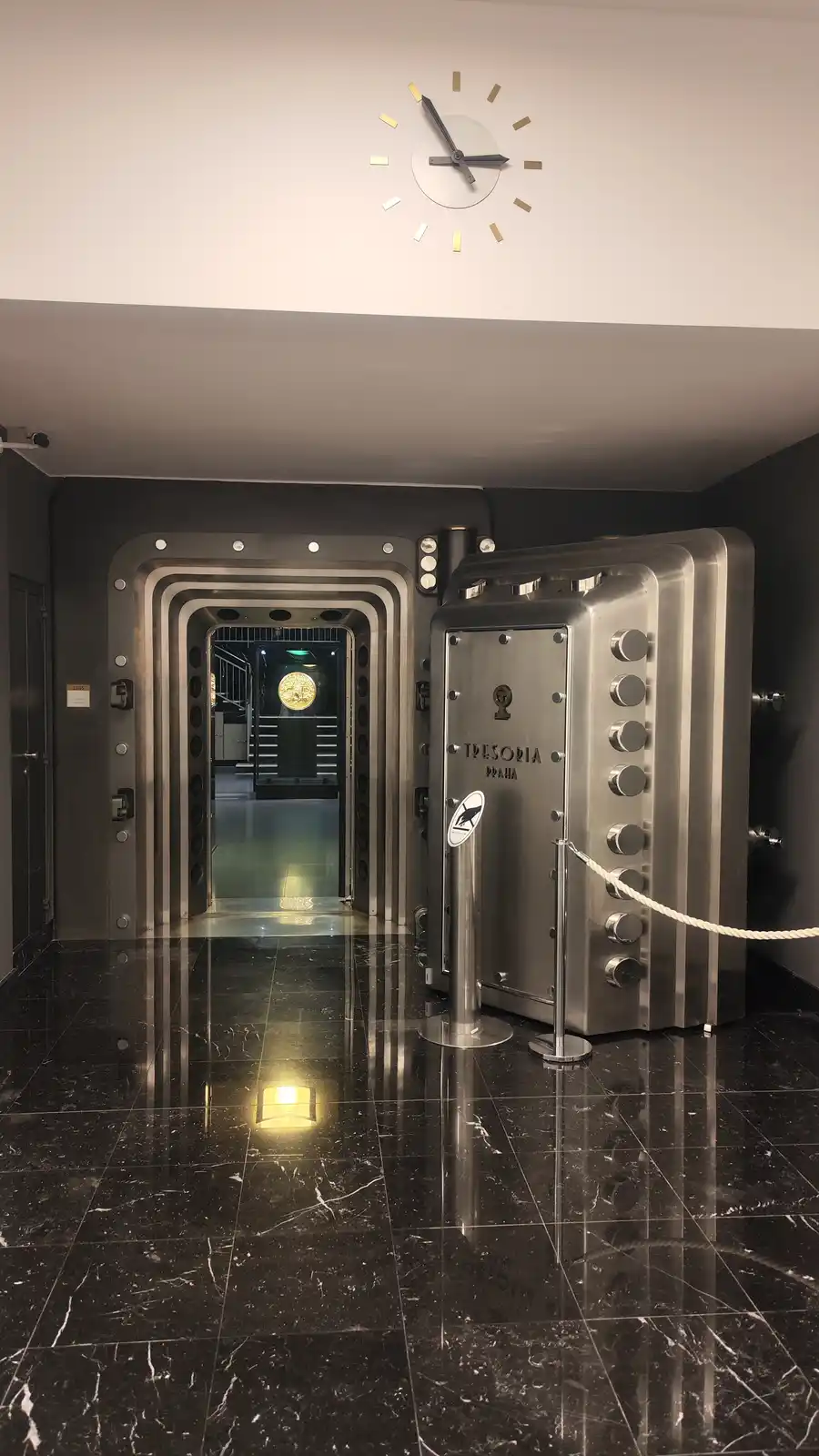 A photo of a large, open, stainless steel vault door labeled 'TRESORIA PRAHA' in a dark, polished marble lobby, with a…