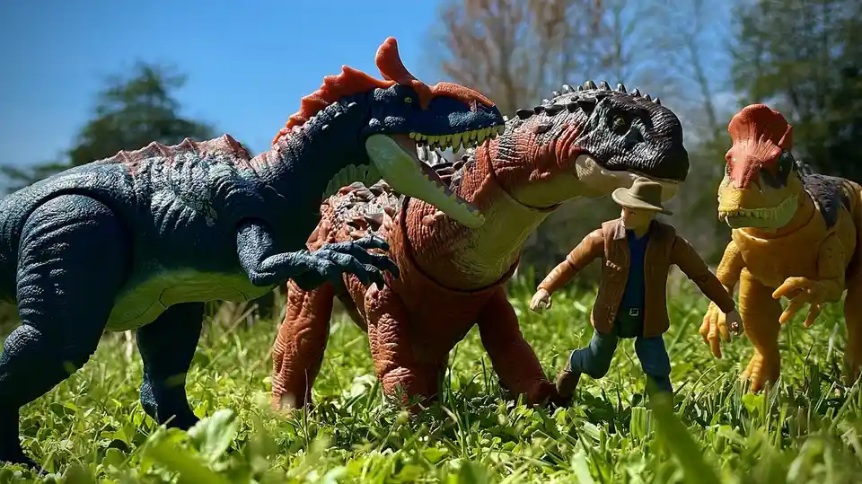 A toy action figure of a man in a hat and vest is running through grass, surrounded by three large, menacing dinosaur toys…
