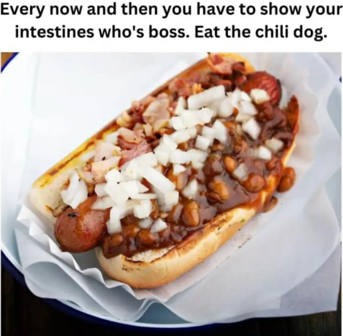 A close-up photo of a chili dog in a bun, topped with chili, beans, bacon, and chopped onions, resting on white paper in a…