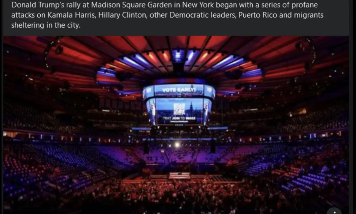 A wide-angle shot of a large, dimly lit arena with tiered seating filled with a crowd, illuminated by red and blue stage…