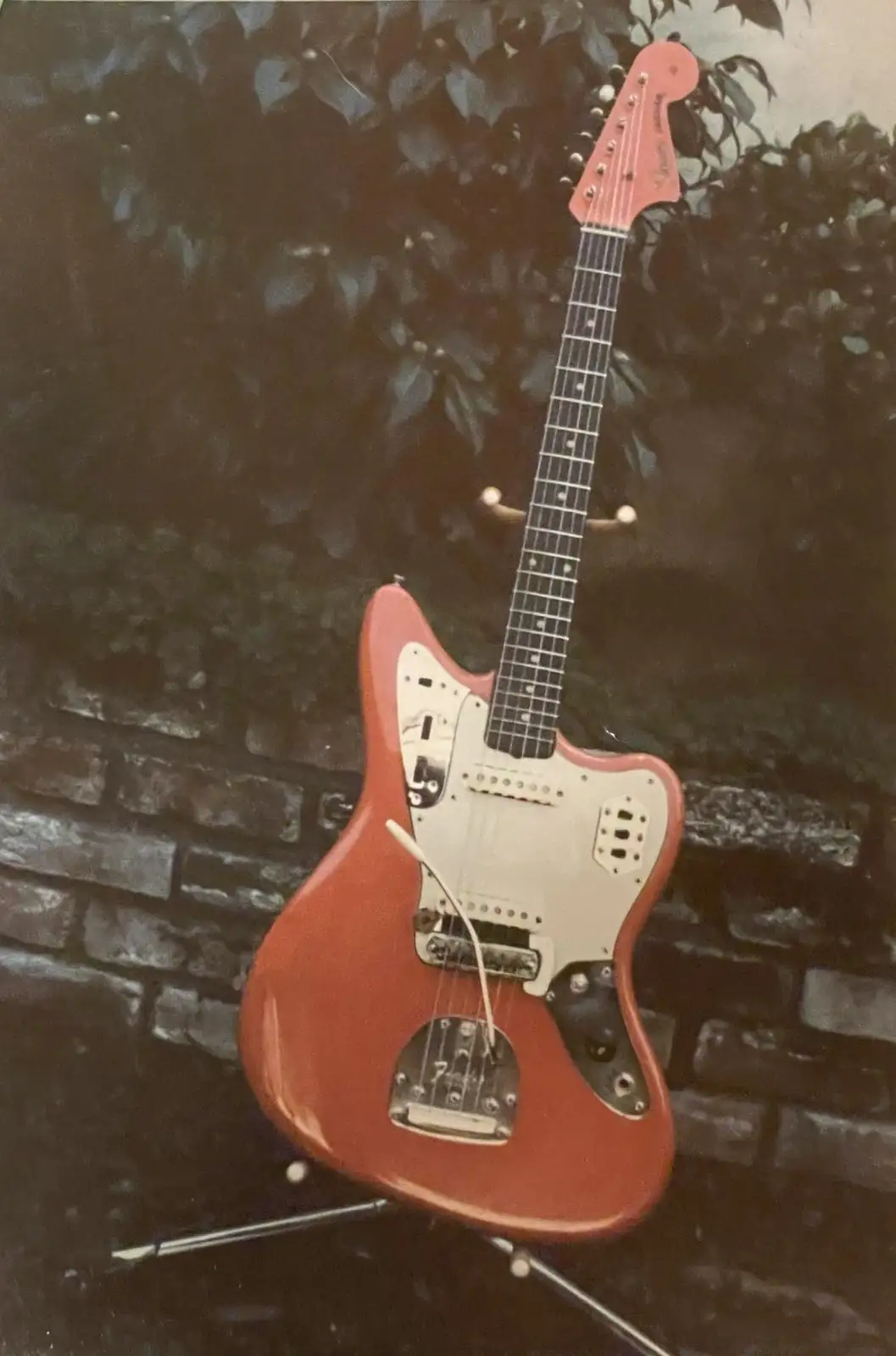 A vintage red Fender Jaguar electric guitar is propped upright on a stand against a dark brick wall with foliage in the…