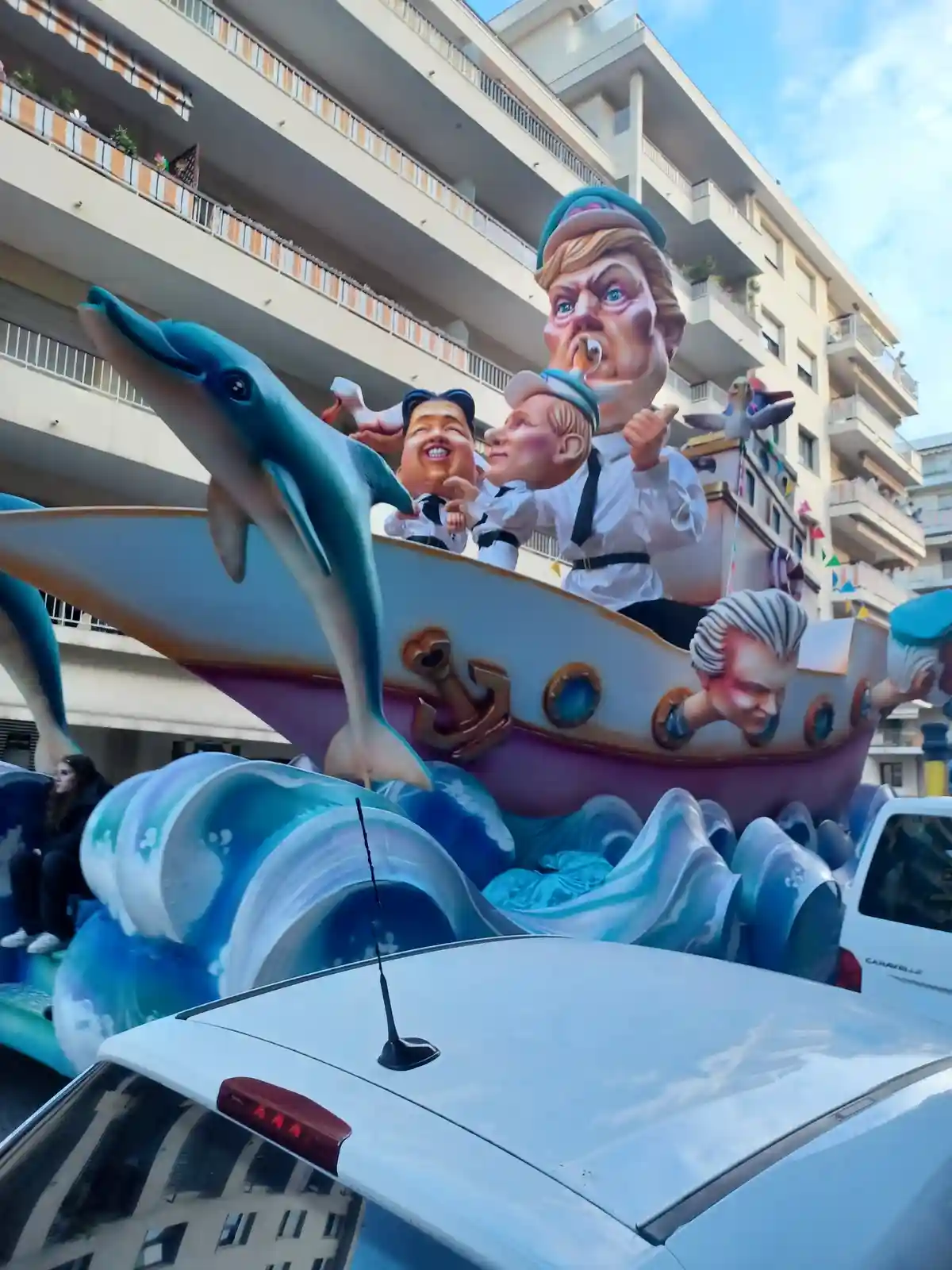 A large, colorful parade float depicts oversized caricatures of political figures on a boat with dolphins and waves. One…