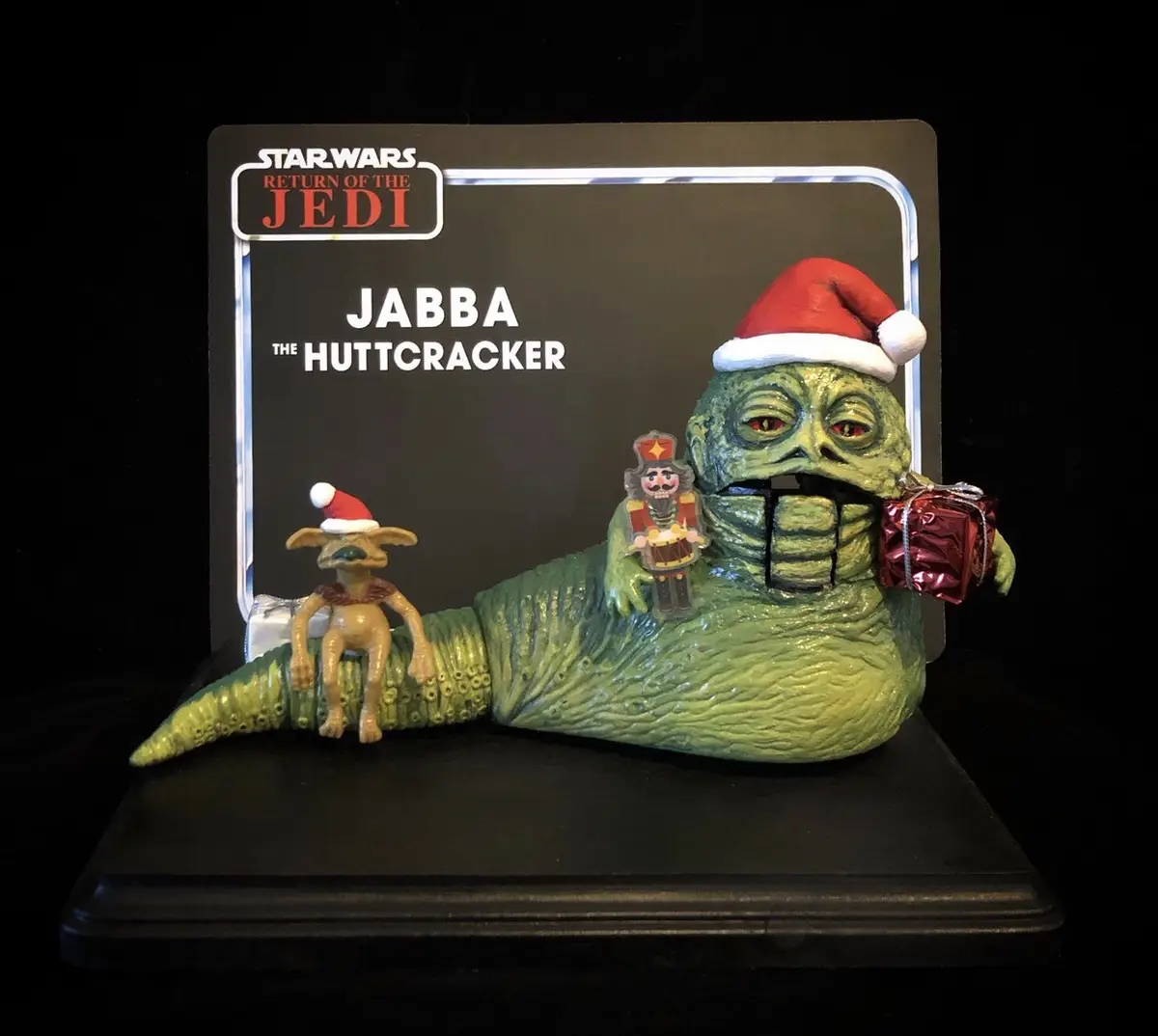 A Star Wars collectible figurine of Jabba the Hutt wearing a Santa hat and holding a wrapped gift, with a small creature…