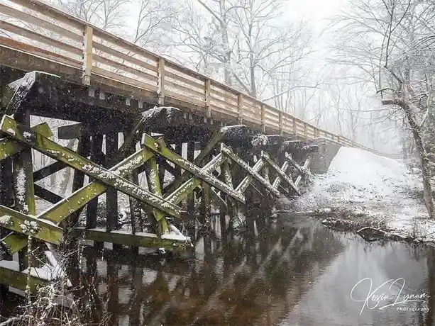 A wooden pedestrian bridge over a river is covered in snow during a snowy day, with bare trees in the background. The…