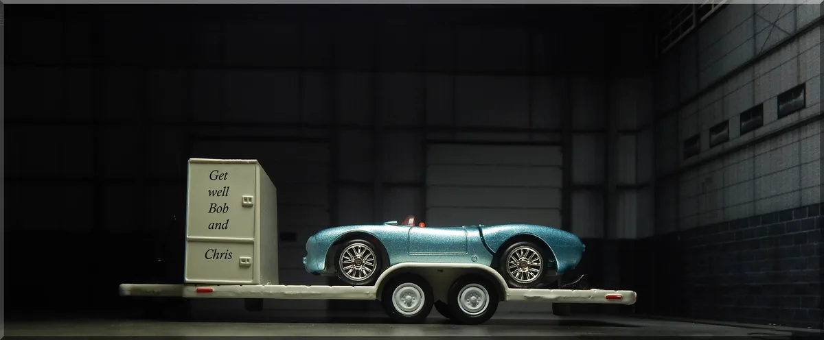 A toy model of a light blue classic sports car is on a white flatbed trailer, which has a beige box attached with the text…
