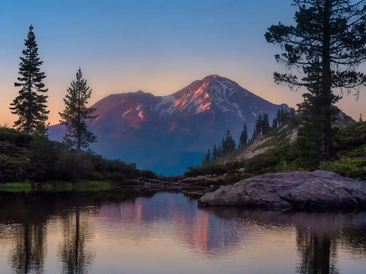 A serene mountain landscape at sunrise or sunset, featuring a snow-capped peak reflecting in a calm lake, framed by tall…