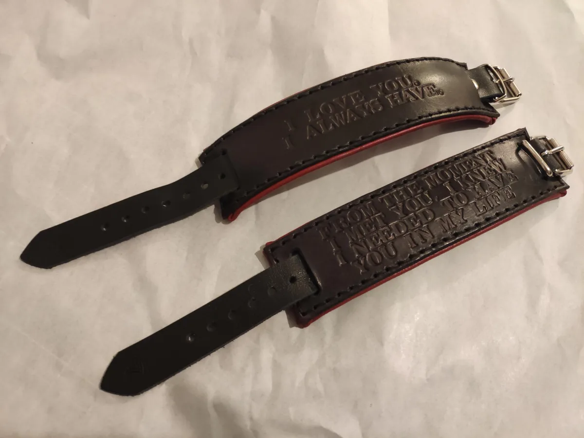 Two dark brown leather bracelets with red inner lining lie on a white crumpled surface. One bracelet reads 'I LOVE YOU. I…