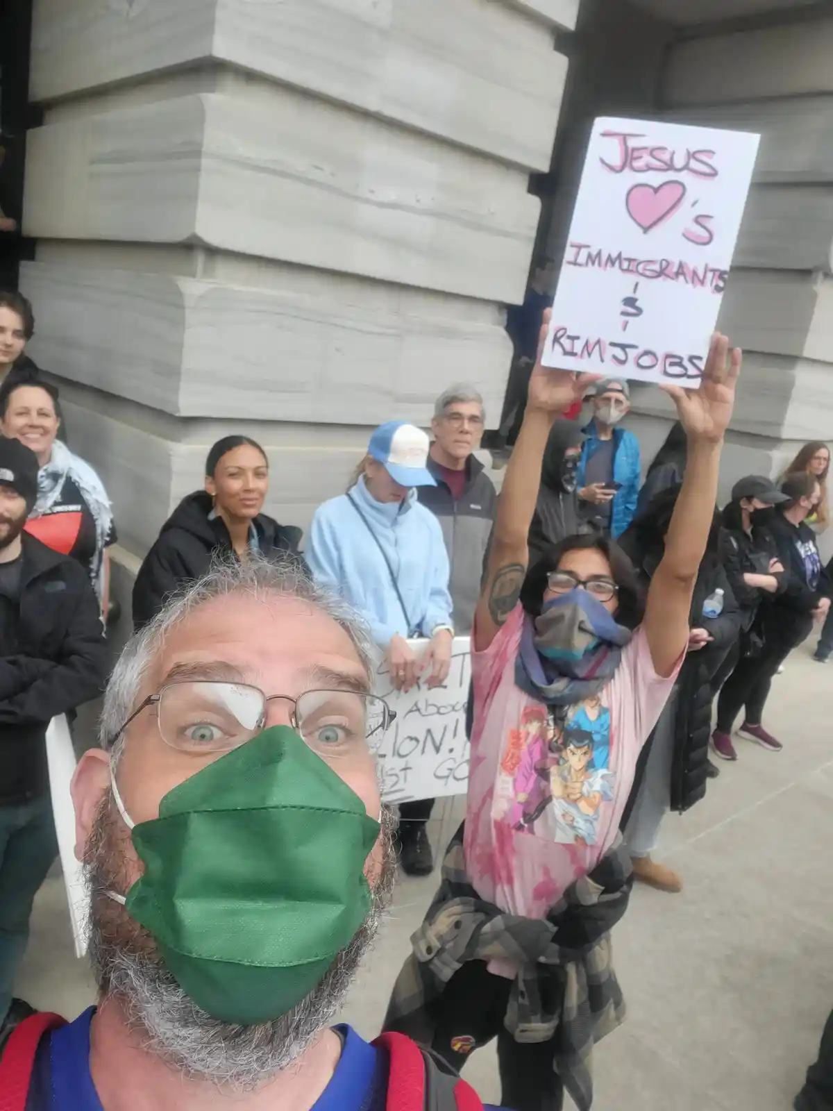 A selfie of a man with a green face mask and glasses, taken outdoors in front of a crowd. Behind him, a person holds a…