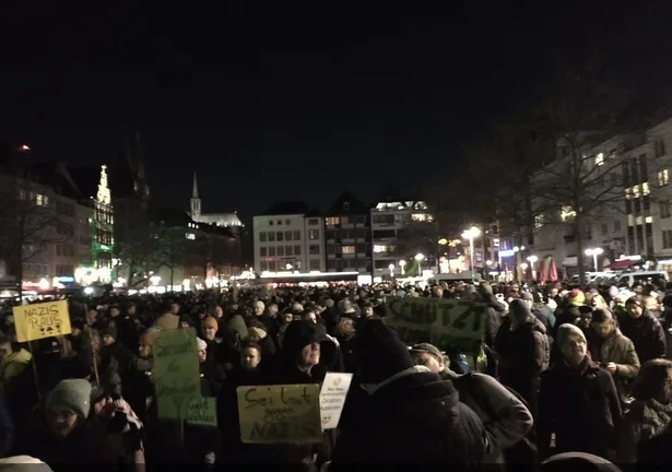 A nighttime photo of a large crowd of people gathered in a city square, holding protest signs. One prominent sign in the…