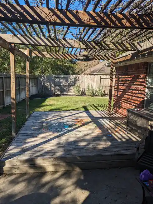 A backyard patio under a wooden pergola with string lights and a hanging fan, featuring a patterned rug on a wooden deck…