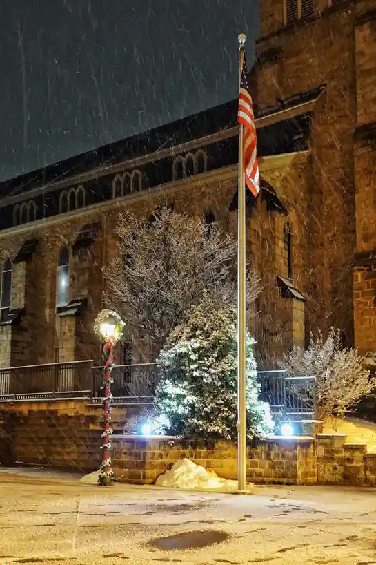 A nighttime photo shows heavy snow falling over a stone building with arched windows, a lit-up Christmas tree, and a…