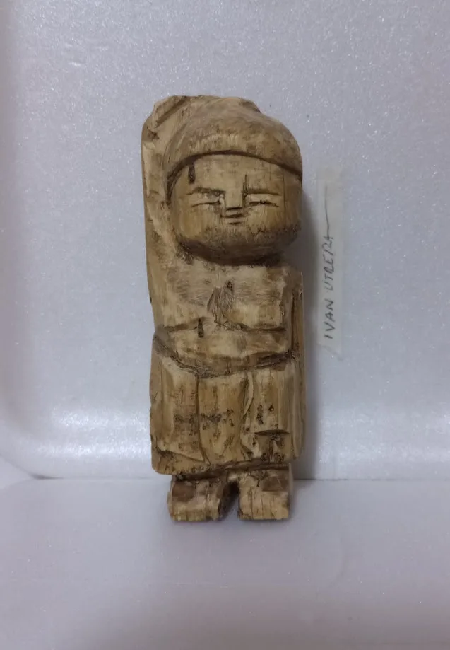 A crudely carved wooden figurine with a simple face and draped clothing stands upright on a white surface, next to a label…
