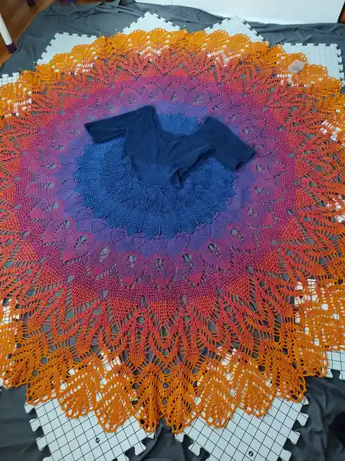 A large, circular, hand-crocheted shawl with a vibrant gradient from orange to purple is spread out on a grey fabric and…