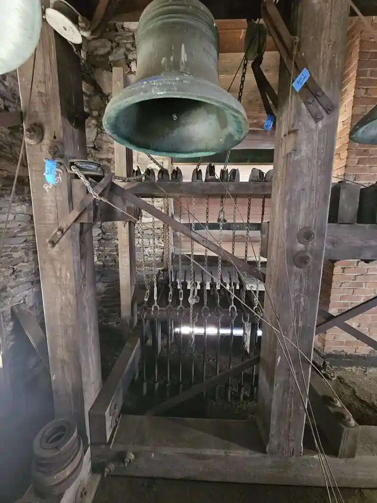 A large, weathered bronze bell hangs inside a wooden bell tower structure, suspended by chains and ropes attached to a…