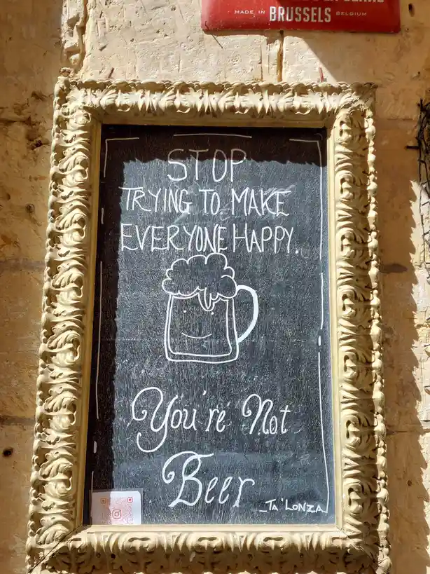 A chalkboard sign in an ornate gold frame mounted on a stone wall reads 'STOP TRYING TO MAKE EVERYONE HAPPY. You're Not…