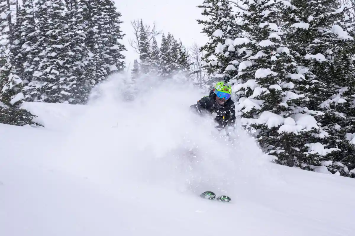 A skier in a bright green helmet and blue goggles carves through deep powder snow, kicking up a large cloud of snow as they…