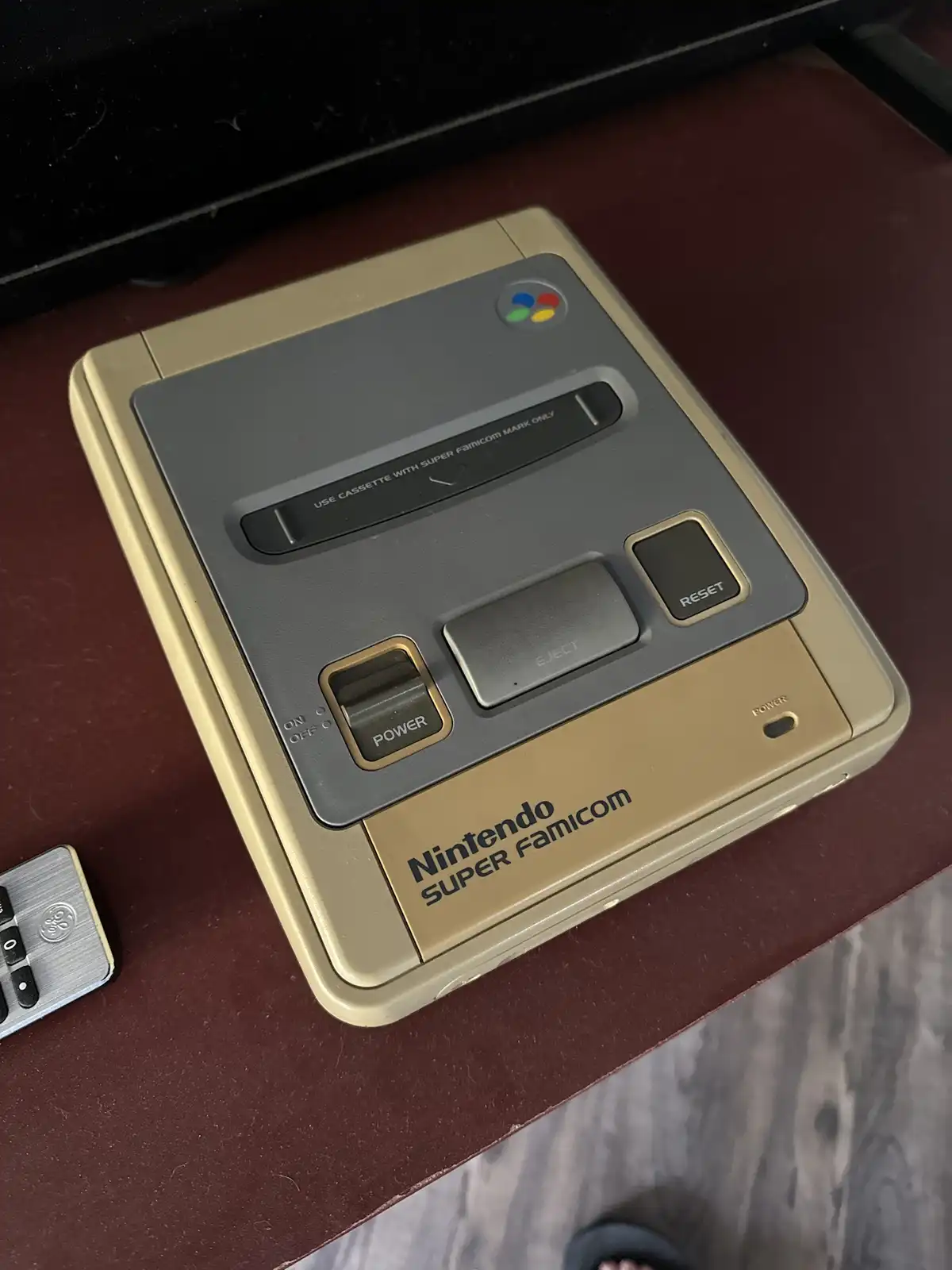 A close-up photo of a beige Nintendo Super Famicom console sitting on a dark red surface, showing its power, eject, and…