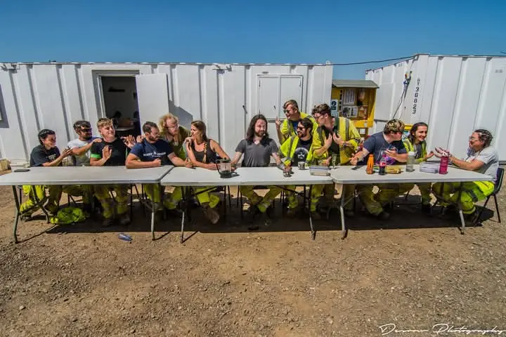 A group of people, many wearing yellow high-visibility clothing, are seated around long tables outdoors in front of white…