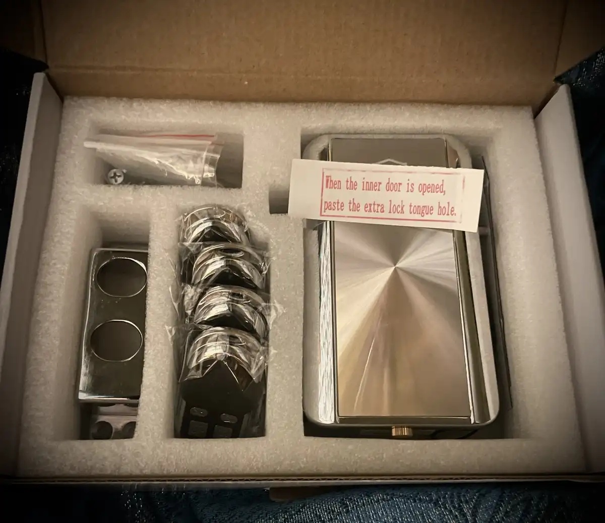 An open cardboard box contains a new door lock set with foam padding, including a large brushed metal lock body, a metal…