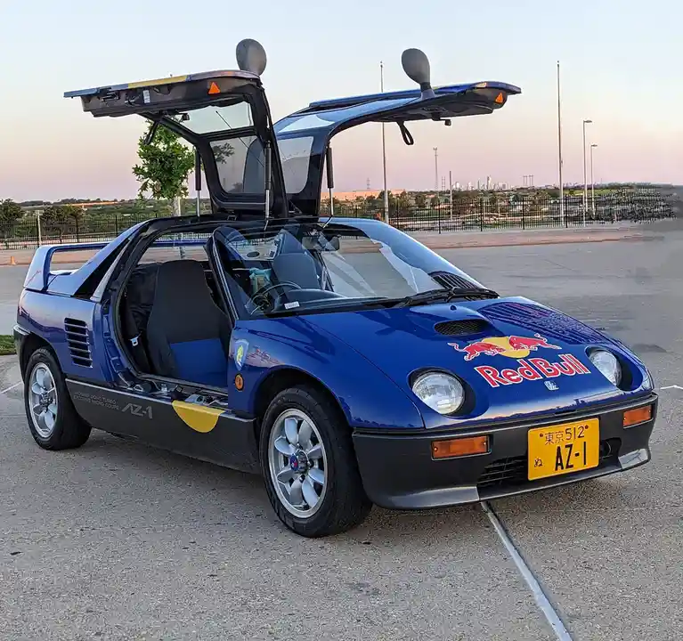 A blue Honda AZ-1 sports car with its gull-wing doors open, parked in a lot with a city skyline in the background. The car…