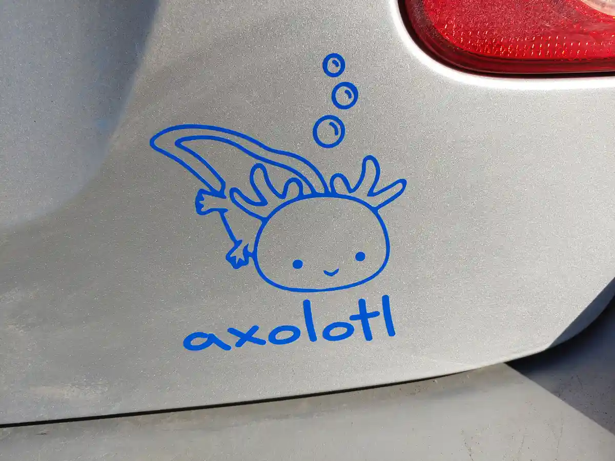 A blue cartoon axolotl sticker with bubbles above its head is affixed to the rear side of a silver car, near the red…