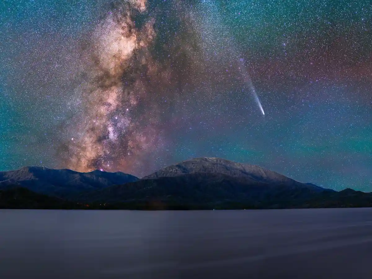 A stunning night sky photograph shows the Milky Way galaxy arching across a star-filled teal and purple sky, with a bright…
