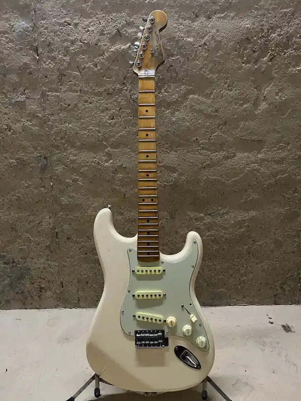 A cream-colored Fender Stratocaster electric guitar stands upright on a stand against a rough concrete wall. The guitar has…