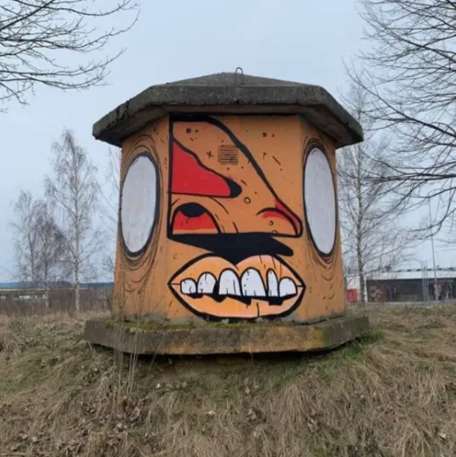a concrete structure, likely an old utility or military bunker, is painted with a cartoonish face featuring large white…