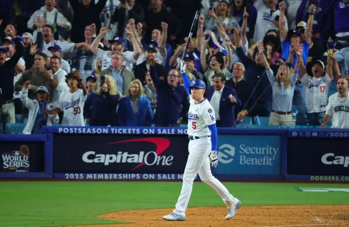 a baseball player in a white dodgers uniform with number 5 raises his bat triumphantly on a field, while a crowd of excited…