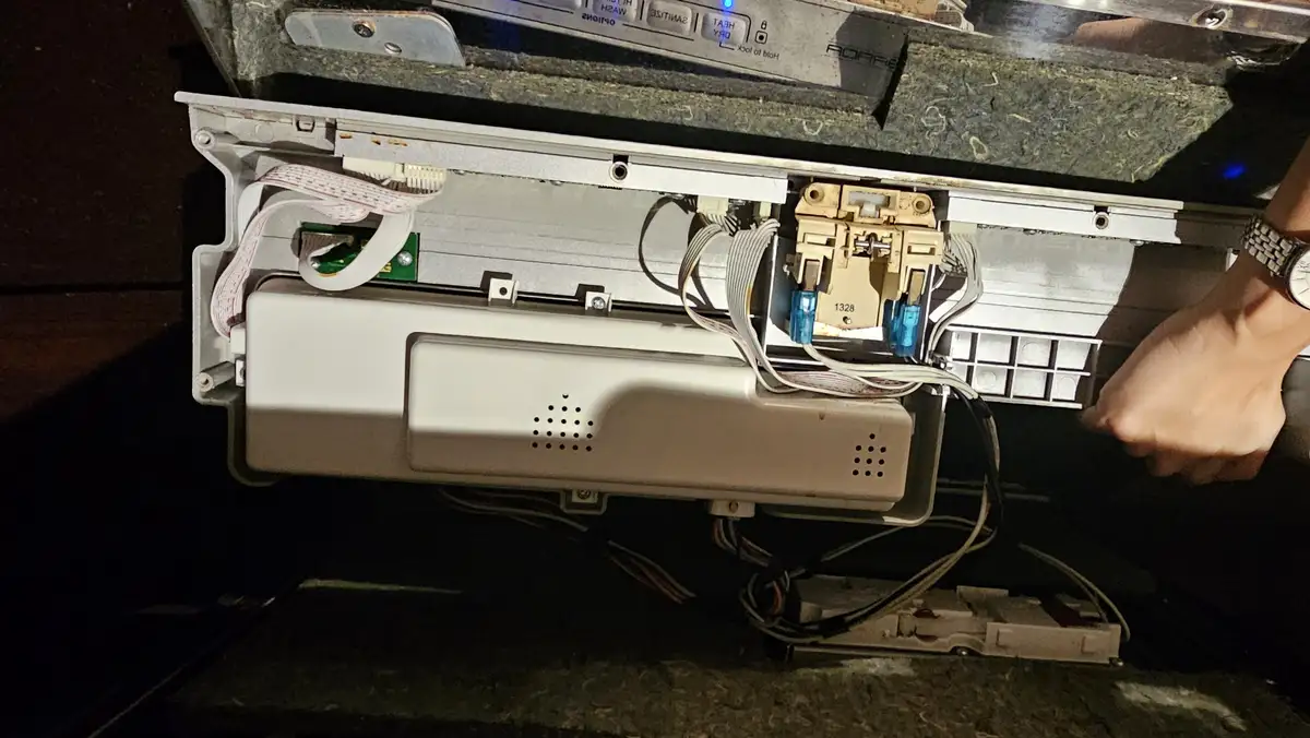 A close-up photo shows the internal wiring and components of a disassembled appliance, likely a dishwasher, with a person's…