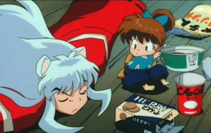 In this anime still, a young girl sits on the floor surrounded by empty snack and drink containers, looking concerned as she…