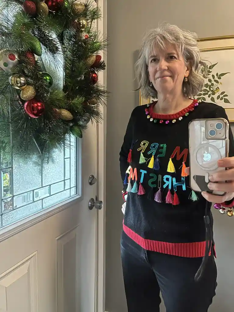 A woman with gray hair takes a selfie in front of a decorated door, wearing a black sweater with the words 'MERRY CHRISTMAS'…