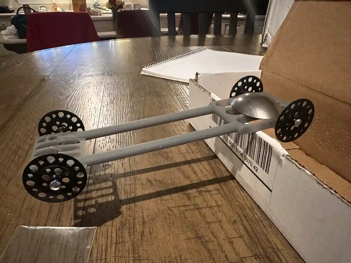 A 3D-printed car chassis with black perforated wheels and a metal spoon serving as the front hood sits on a wooden table…