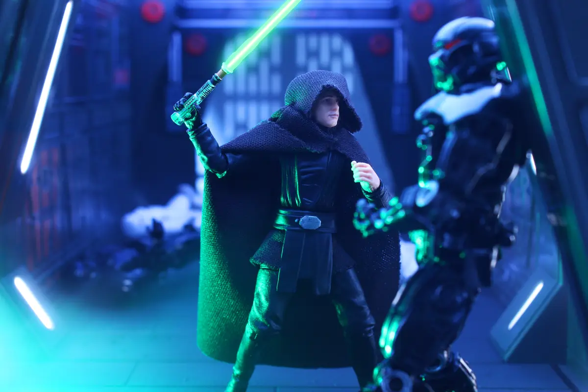 A Star Wars action figure of a hooded, dark-cloaked character holding a green lightsaber stands in a sci-fi corridor, facing…