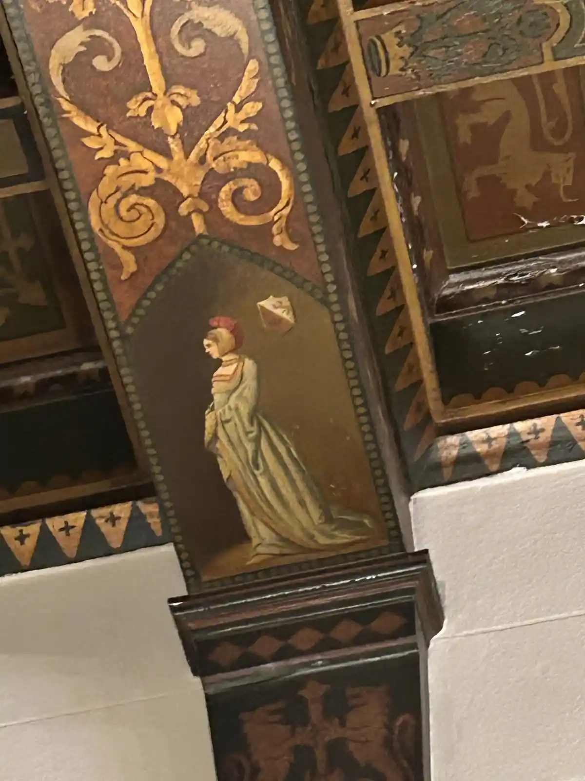 A close-up of ornate, aged ceiling or wall paneling with a painted medieval-style woman in a long gown and red headdress…